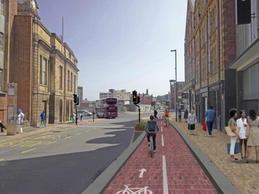 A street scene showing a segregated red cycle lane running past shops and buildings in an urban area. People are walking on the pavement to the right, and cyclists are using the marked cycle lane. Traffic lights and a bus are visible on the road to the left, with more buildings and the city skyline in the background.