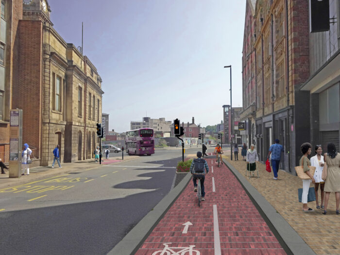 A street scene showing a segregated red cycle lane running past shops and buildings in an urban area. People are walking on the pavement to the right, and cyclists are using the marked cycle lane. Traffic lights and a bus are visible on the road to the left, with more buildings and the city skyline in the background.