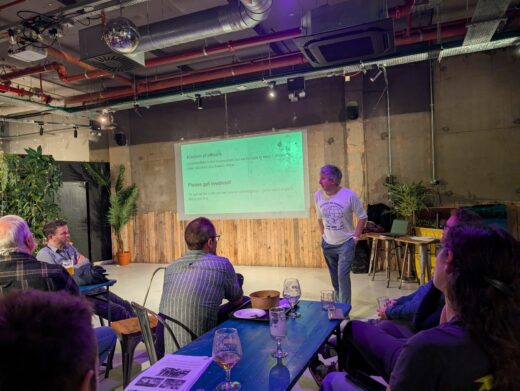A man stands at the front of a room giving a presentation to a seated audience. A slide projected on the wall behind him reads “Election of officers” and “Please get involved!” The setting is informal, with wooden panels, exposed pipes, plants, and drinks on the tables, creating a relaxed community meeting atmosphere.
