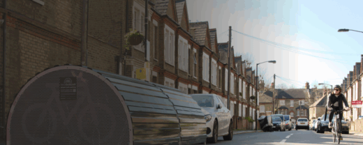 A quiet residential street lined with terraced houses. In the foreground, a metal bike hangar sits by the curb. Cars are parked along the street, and a cyclist wearing a helmet rides toward the camera under a clear sky.